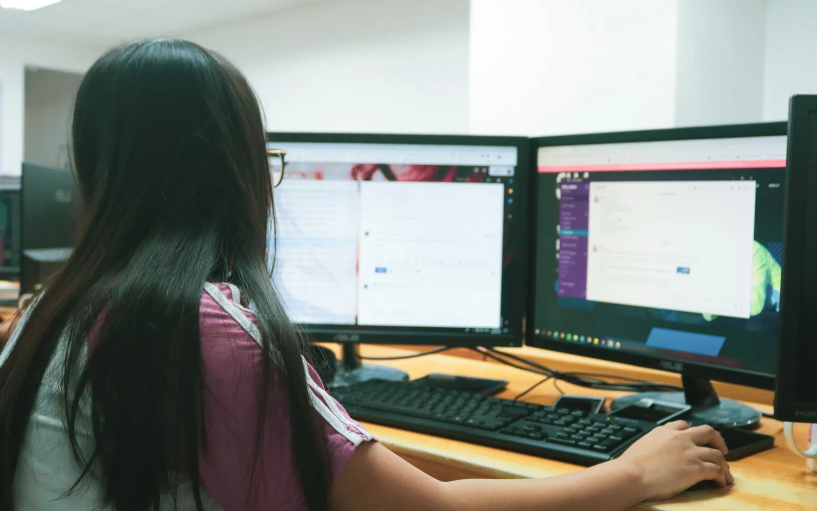 A person seen from behind types at a keyboard while working across two desktop monitors in an office.