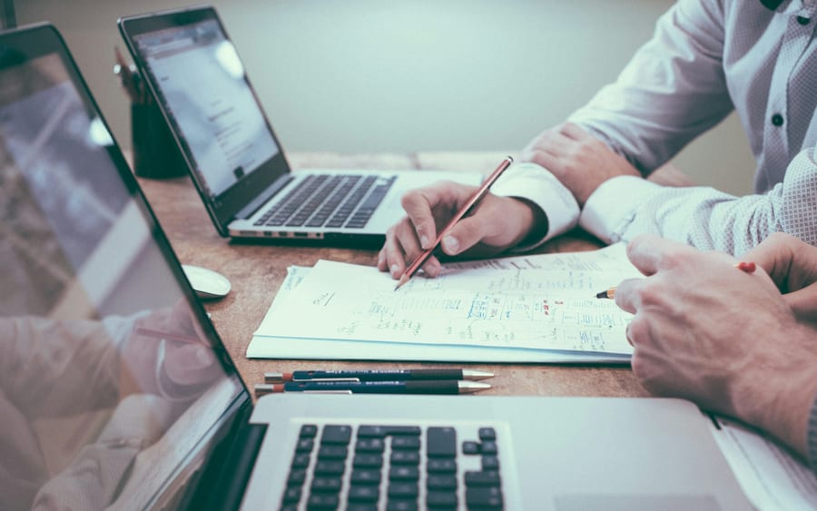 A person writes notes on paper at a desk beside an open laptop while preparing for a meeting.