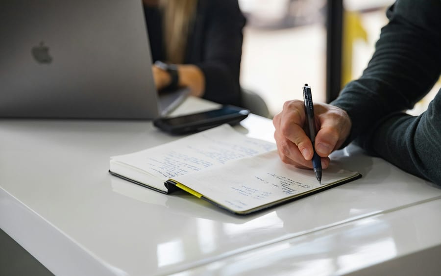 A person writes in a notebook with a pen at a white desk, with a laptop and phone beside it.