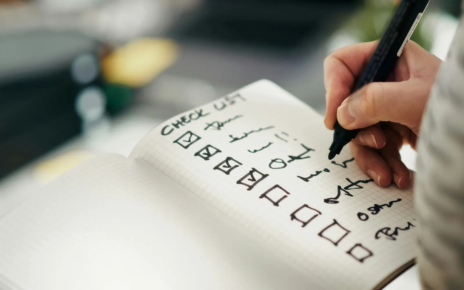 A hand holding a black pen ticks boxes on a handwritten checklist in an open notebook on a desk.